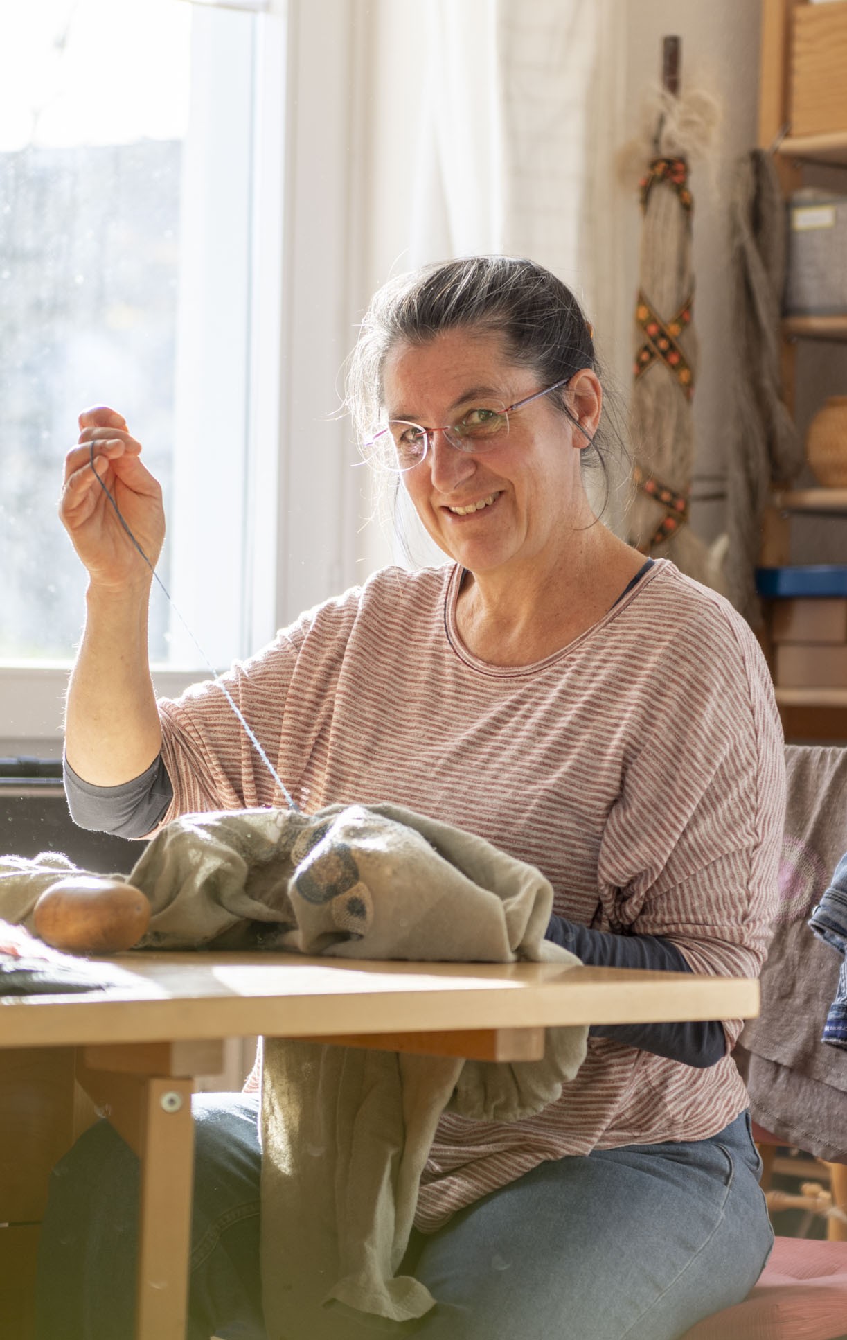 Katja Frefel, die Handwerkerin, sitzt mit geflochteten Haaren am Arbeitstisch und lächelt dem Zuschauenden zu, während sie an einem Projekt weiternäht.